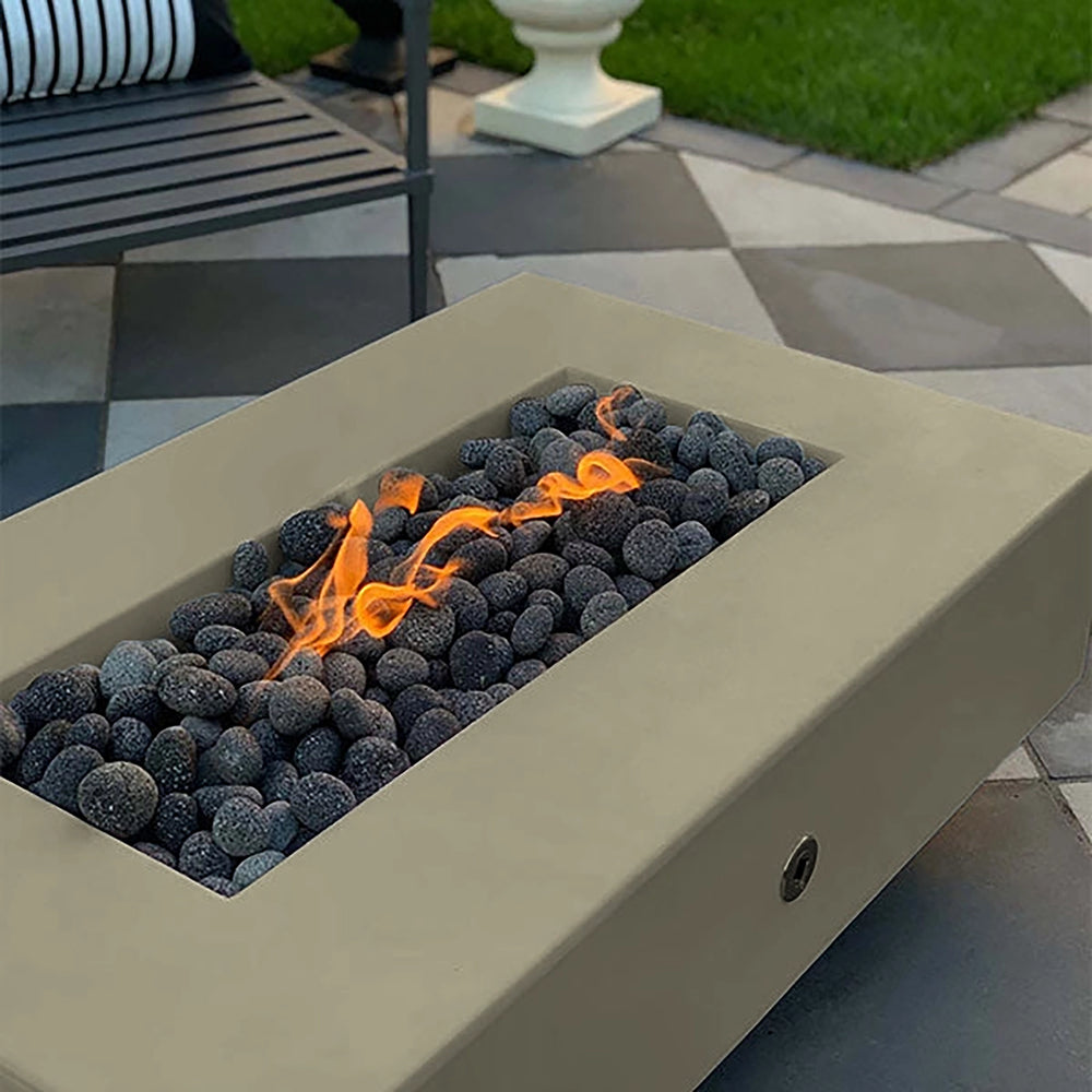 The Outdoor Plus 60-inch Del Mar Concrete Gas Fire Pit in a neutral ash tone, featuring a close-up view with vibrant flames rising through dark lava rocks. Ideal for outdoor ambiance, this rectangular fire pit is designed to enhance backyard settings with a modern, natural gas-fueled flame.