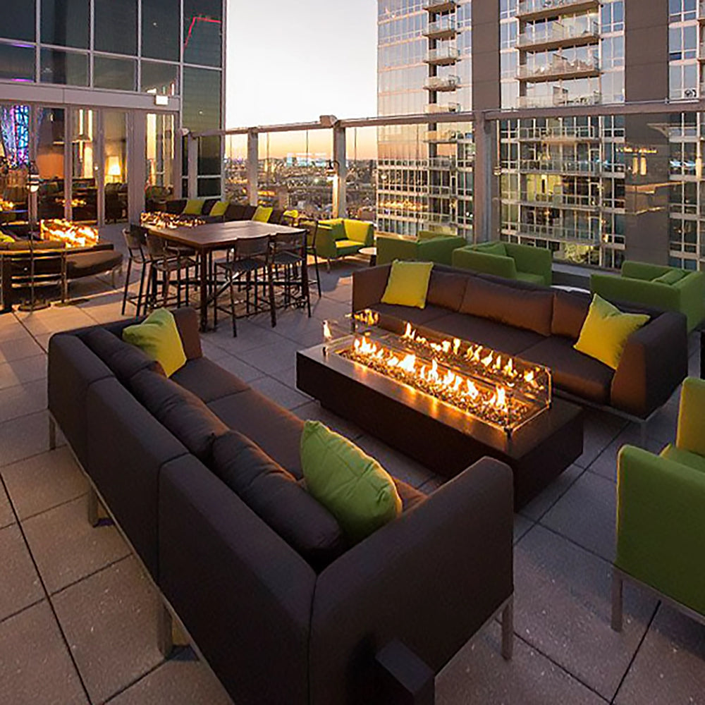 The Outdoor Plus 72-inch Del Mar Concrete Gas Fire Pit elegantly complements an upscale rooftop lounge against a cityscape at dusk. This rectangular concrete fire pit emits a cozy flame, making it ideal for outdoor social spaces. Designed for both luxury residential and commercial environments, this gas fire pit enhances any outdoor area with its modern appeal.