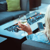 Close-up of The Outdoor Plus 84-inch Del Mar concrete gas fire pit in black, with flickering flames in a bed of fire glass. The scene features a modern outdoor setup with a woman enjoying wine, showcasing luxury and relaxation