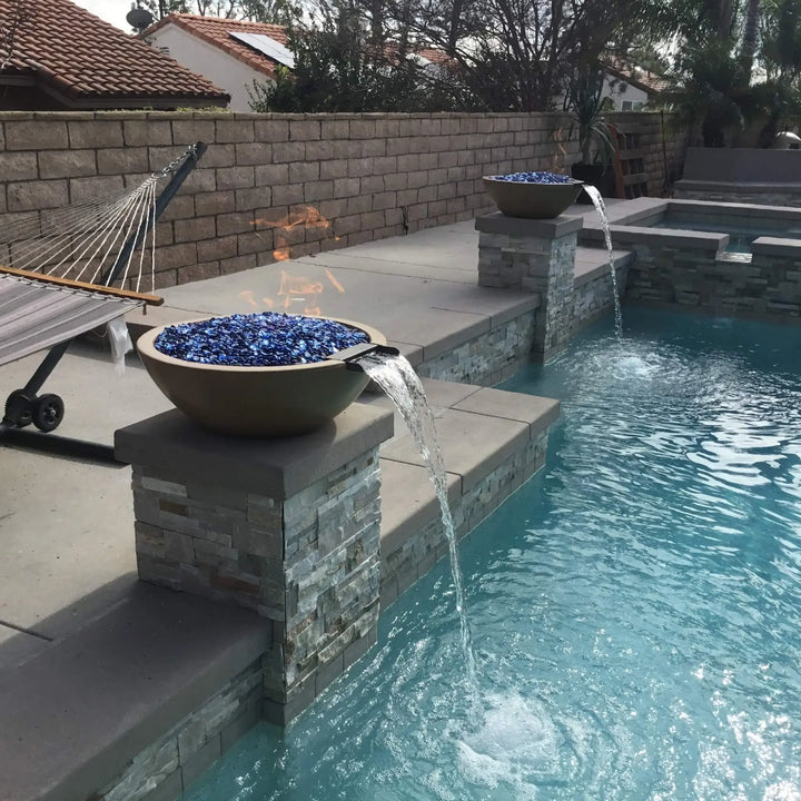 A serene outdoor pool setup featuring two Sedona Round Fire and Water Bowls by The Outdoor Plus in a smooth, earthy concrete finish. Each bowl emits a soft flame over deep blue fire glass while water gently cascades into the pool below. A nearby hammock and modern stonework create a peaceful backyard retreat perfect for relaxation and warmth.