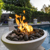 A close-up of The Outdoor Plus Sedona Round Fire Bowl in a light concrete finish, filled with natural lava rocks and bright orange flames dancing at sunset. Positioned beside a modern pool and surrounded by manicured landscaping, this design brings warmth, elegance, and ambiance to any luxury outdoor space.