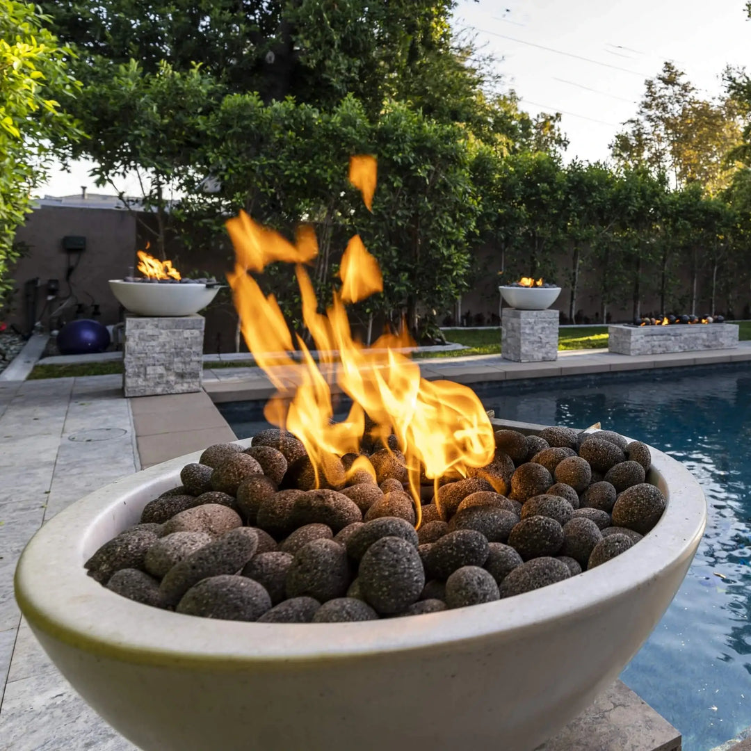 A close-up of The Outdoor Plus Sedona Round Fire Bowl in a light concrete finish, filled with natural lava rocks and bright orange flames dancing at sunset. Positioned beside a modern pool and surrounded by manicured landscaping, this design brings warmth, elegance, and ambiance to any luxury outdoor space.