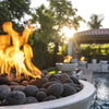 A glowing Sedona Round Fire Bowl from The Outdoor Plus, filled with lava rocks and set aglow by the setting sun. Positioned poolside with other fire features in the background, the light-colored concrete bowl brings a cozy, modern vibe to this beautifully landscaped outdoor entertainment area.
