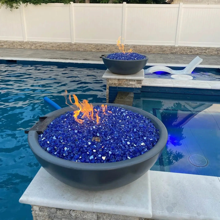 A sleek and modern backyard poolscape featuring The Outdoor Plus Sedona Round Fire and Water Bowl in a dark concrete finish. Filled with vibrant cobalt blue fire glass, the bowl showcases a lively flame as it rests on a stone pillar beside a tranquil pool. The deep blue water and modern lounge chairs enhance this luxurious and relaxing outdoor setting.