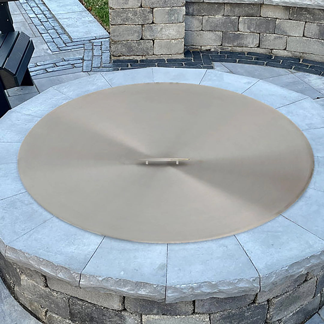 The Outdoor Plus Round Stainless Steel Burner Cover in a lifestyle setting, placed over a built-in fire pit surrounded by a stone patio. The 23-inch round burner cover is made from high-quality brushed stainless steel and is designed to protect the fire pit from debris and harsh weather elements. Ideal for outdoor patio settings, this cover blends seamlessly with its surroundings while maintaining a polished and modern look