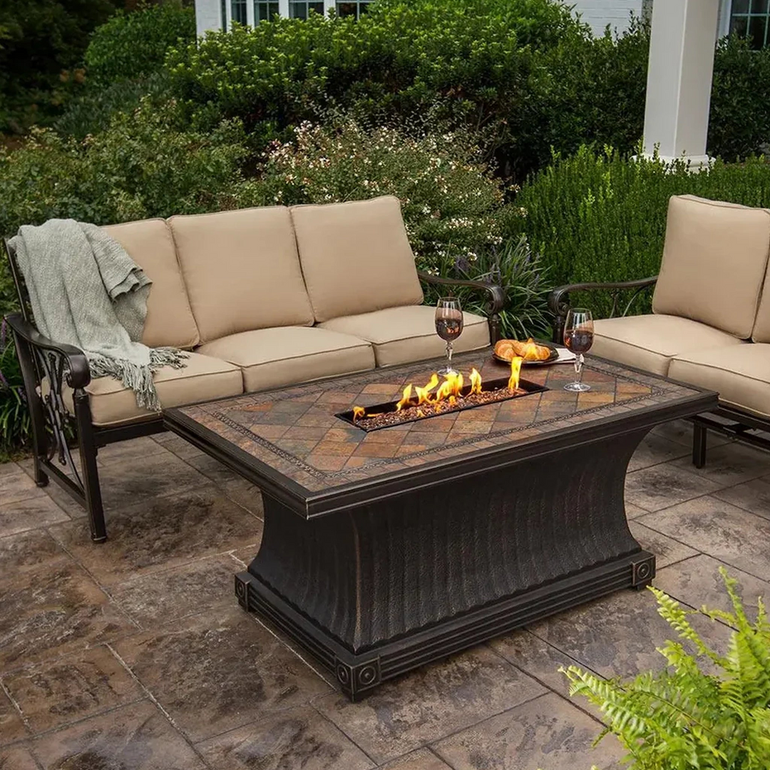 Cozy outdoor seating area featuring a rectangular fire pit table with a decorative stone top, surrounded by beige cushioned sofa and chairs on a stone patio, set against a backdrop of lush greenery