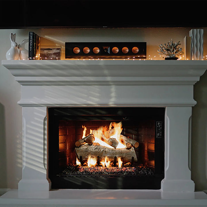 A cozy living room scene featuring a white fireplace mantel with vented gas logs from the Mountain Birch collection by Real Fyre. The fireplace is ignited, showcasing the realistic birch logs arranged in a traditional wood stack formation, with warm flames glowing through. The logs' detailed bark texture and natural white birch appearance add a rustic touch to the modern, clean fireplace setting. Decorative elements sit atop the mantel, creating a warm, inviting ambiance ideal for living spaces.