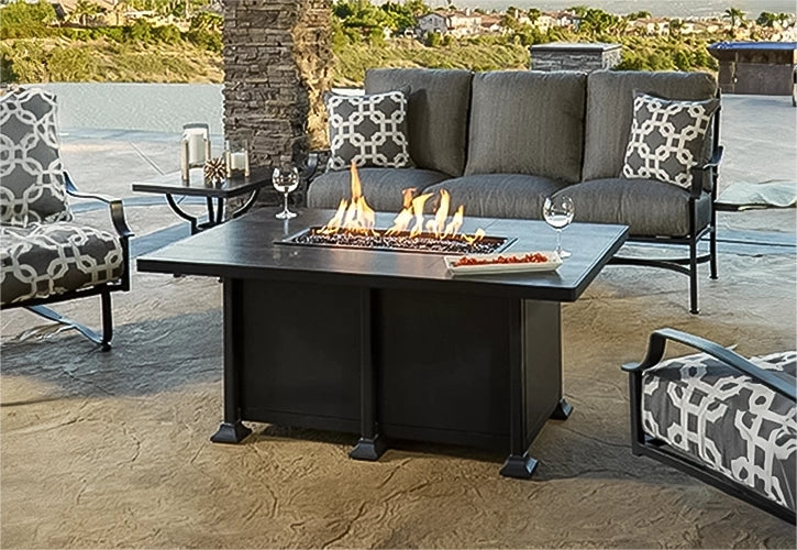 Cozy outdoor seating area featuring a rectangular black fire pit table, gray sofa, and chairs with patterned cushions, set against a scenic background with wine glasses on the table